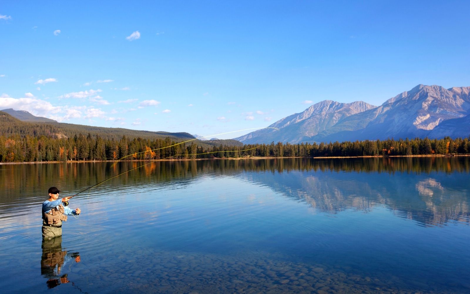 A man wearing waders casts his fly fishing pole in a lake