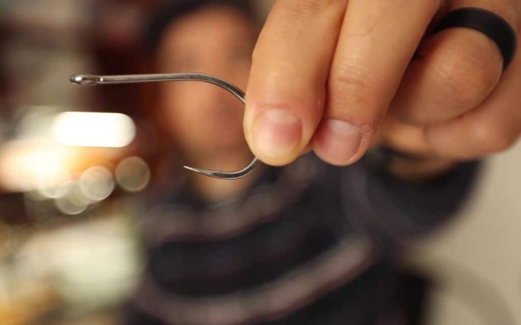 An angler is holding a large barbless fly fishing hook on a blurry background.