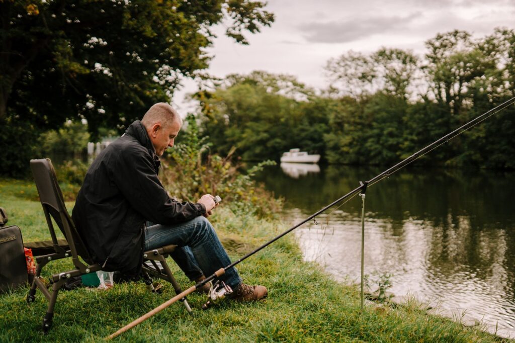 An angler seated in a chair on a riverbank checks his phone while his rod sits in a holder