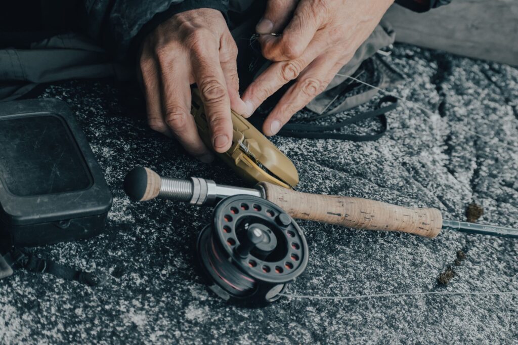 Angler's hands working with a fly reel and rod, preparing leader and line on a rocky surface