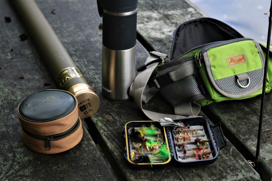 Open fly box showing a variety of fly fishing fly patterns beside a rod and fishing vest