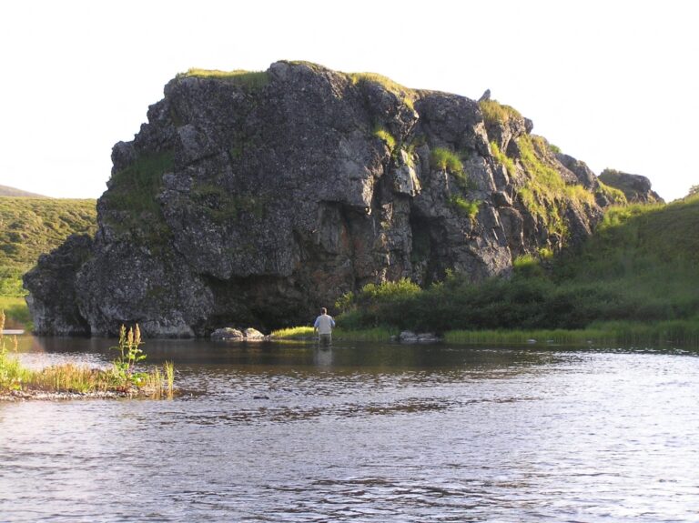 An angler fishing in a broad river beside rocky banks